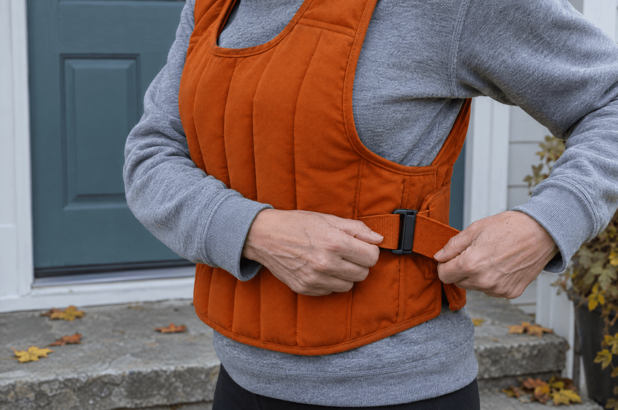 Hands fasten the side strap of an orange weighted vest on a front step on an autumn morning.